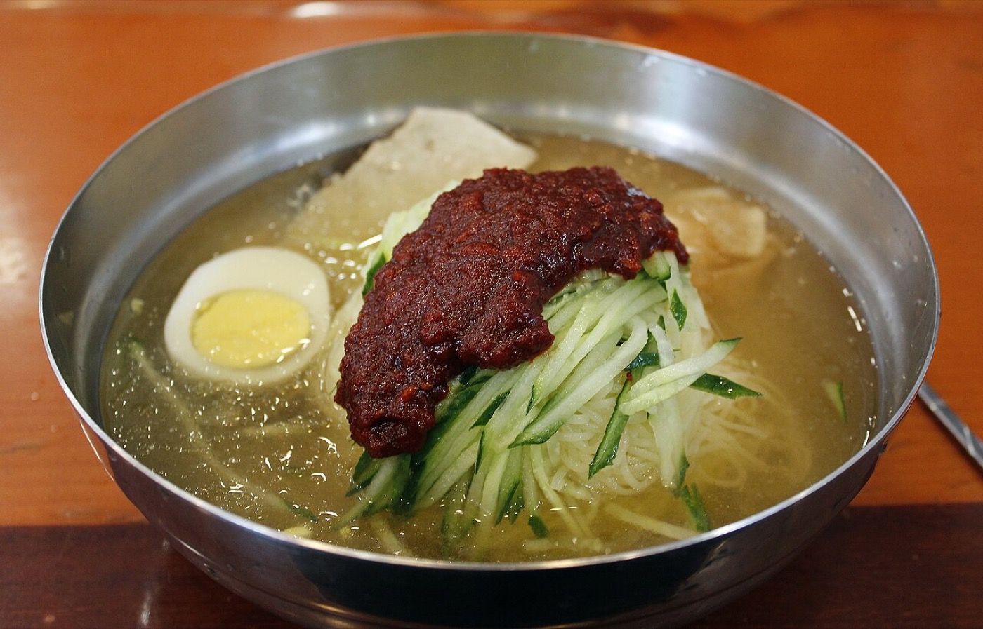 부산 밀면 한 그릇이에요. 스테인리스 대접에 맑은 육수가 담겼고, 가느다란 면 위에 채 썬 오이와 삶은 달걀 반쪽, 무가 놓였어요. 가운데에 붉은 양념장이 한 덩이 얹혀 있어요.