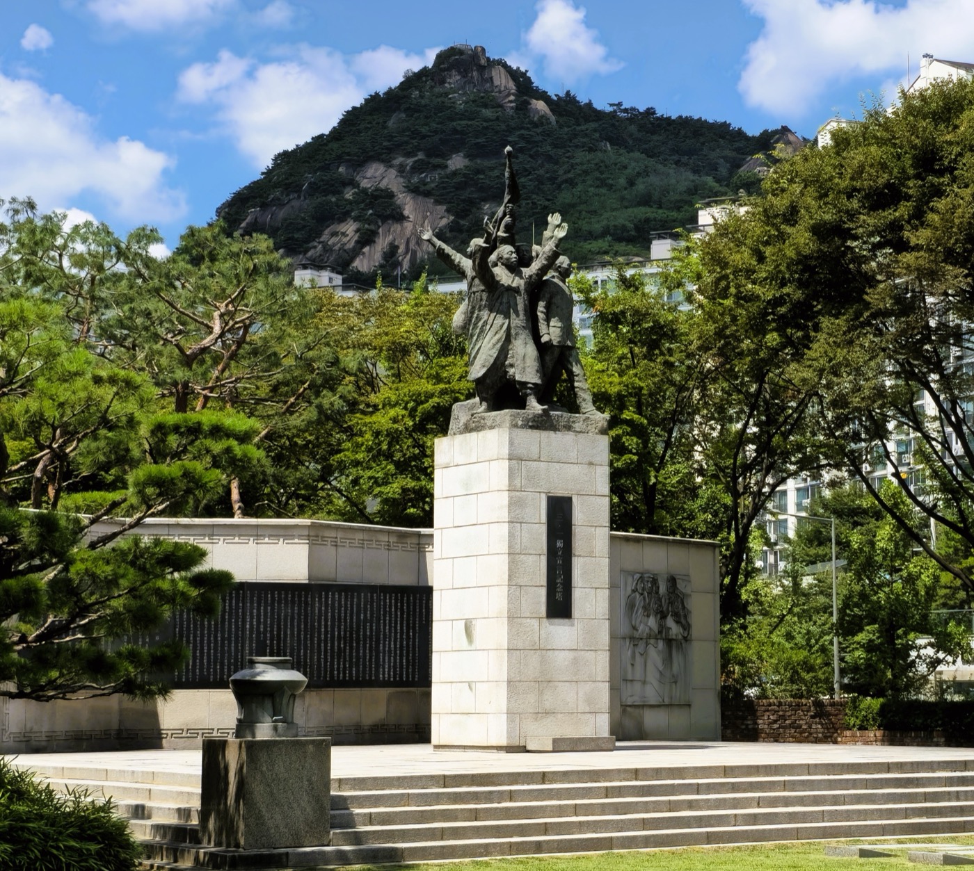 서울 서대문독립공원에 있는 3·1독립선언 기념탑이에요. 네모난 돌 받침대 위에 팔을 높이 치켜들고 깃발을 든 사람들의 청동상이 서 있어요. 받침대 앞 검은 판에는 「三·一 獨立宣言記念塔(삼일 독립 선언 기념탑)」이라고 적혀 있어요. 탑 왼쪽 뒤 낮은 검은 벽에는 독립선언서가 빽빽하게 새겨져 있고, 오른쪽 벽에는 사람들을 새긴 조각이 있어요. 뒤로 바위산이 보여요.