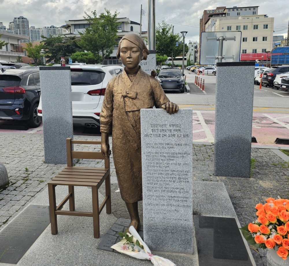전북 익산역 앞 광장에 세워진 평화의 소녀상이에요. 한복을 입은 단발머리 소녀가 서서, 한 손을 글이 새겨진 돌비석 위에 얹고 있어요. 어깨에는 작은 새가 앉아 있어요. 옆에는 나무 의자 하나가 비어 있고, 발밑에 꽃다발이 놓여 있어요.